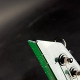 Close-up of the corner of a vintage electric guitar headstock, showing a green-painted edge, a white top, and part of the tuning pegs against a dark background.