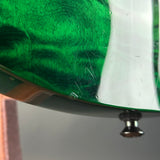 Close-up of a glossy green guitar body with a white edge and a visible metal tuning peg below, photographed against a blurred gray background.