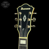Close-up of an Ibanez electric guitar headstock against a black background, showing the gold Ibanez logo, six tuning pegs, and gold binding.