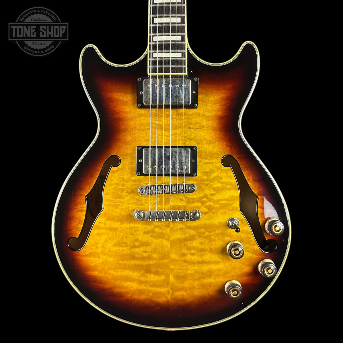 A classic electric guitar with a flamed maple body, featuring a vibrant sunburst finish that transitions from bright yellow-orange in the center to dark brown and black at the edges. It has black humbucker pickups, chrome hardware, and a white binding edge, shown against a solid black background with a Tone Shop watermark in the top left corner.