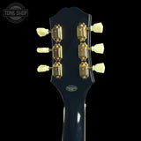 Close-up of the headstock of a dark blue electric guitar against a black background, showing six gold tuning pegs with cream-colored knobs. A Tone Shop watermark is visible in the top left corner.