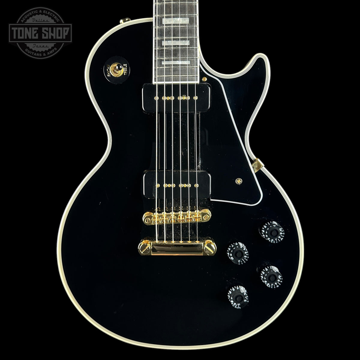 A black electric guitar with white binding and gold hardware, shown from the front against a black background. A Tone Shop watermark is visible in the top-left corner.