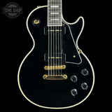 A black electric guitar with white binding and gold hardware, shown from the front against a black background. A Tone Shop watermark is visible in the top-left corner.