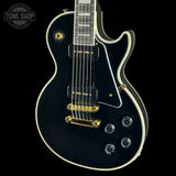 A black electric guitar with gold hardware and white binding, shown against a solid black background. A Tone Shop watermark is visible in the top-left corner.