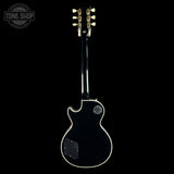 A black electric guitar shown from the back against a solid black background, featuring a glossy finish, white binding, and gold tuning pegs. A circular 'TONE SHOP' watermark is visible in the top-left corner.