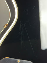 Close-up of a glossy black guitar body with visible scratches, dust, and hair on the surface, along with part of a white pickguard and a gold screw.