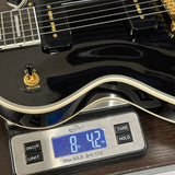 Close-up of a glossy black electric guitar body resting on a digital scale, with the scale display showing the weight in ounces. The guitar has gold hardware, white binding, and a visible Wilbur brand decal near the tailpiece.