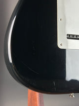 Close-up of the glossy black body of an electric guitar, showing a white pickguard, control hole covers, and a small portion of the wood neck at the right edge.