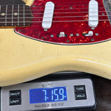 Close-up of a guitar body resting on a digital scale, showing a cream-colored guitar with a red pickguard and white pickups. The scale displays a weight of 7.