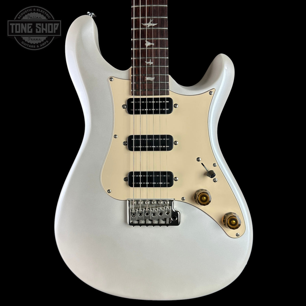 A white electric guitar with a cream-colored pickguard, black pickups, and gold-toned controls, shown against a solid black background. A Tone Shop watermark is visible in the top-left corner.