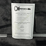 A white paper certificate label for Charvel Custom Shop Custom Select, showing product certification details, photographed on a dark textured fabric background.