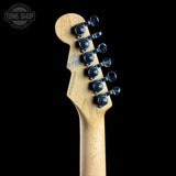 Close-up of the headstock of an electric guitar, showing a natural maple wood finish, six black tuning pegs, and a blue signature on the back of the headstock, set against a solid black background.