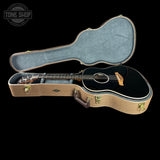 An acoustic guitar with a glossy black body and natural wood headstock, displayed inside an open tan hard case with a gray felt lining, set against a solid black background.