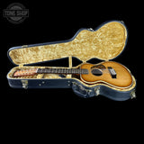 An acoustic guitar with a natural wood finish resting inside an open hard case with a gold marbled interior, shown against a solid black background. A Tone Shop logo watermark is visible in the top-left corner.