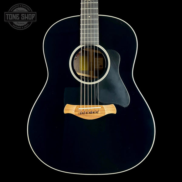 A close-up studio shot of the front of a dark acoustic guitar against a black background, showing the glossy black body, white binding, sound hole, pickguard, and natural wood bridge. A Tone Shop watermark is visible in the upper left corner.