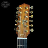 Close-up of a 12-string acoustic guitar headstock in a warm brown wood finish with gold-toned tuning pegs and strings, shown against a solid black background. A Tone Shop watermark is visible in the top-left corner.