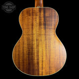 Close-up of the back of an acoustic guitar showing a rich, warm brown wood grain finish on a solid black background, with a Tone Shop logo in the top left corner.