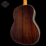 Close-up of the back of an acoustic guitar showing a rich dark brown wood grain finish and white edge trim, set against a solid black background with a Tone Shop watermark in the top left.