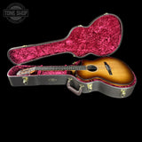 An acoustic guitar with a sunburst finish displayed inside an open hard case with a bright pink velvet interior, set against a solid black background. A Tone Shop watermark is visible in the top-left corner.