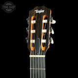 Close-up of the headstock of a Taylor classical guitar against a solid black background, showing the dark wood finish, orange inner soundboard slots, white tuning pegs, and the Taylor logo.