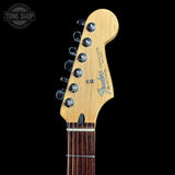 Close-up of a Fender Stratocaster electric guitar headstock in natural maple wood, featuring six silver tuning pegs and the blue Fender logo, set against a solid black background.