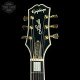 Close-up of the headstock of an Epiphone Lucille Stevens electric guitar against a solid black background, showing the gold tuning pegs, white script logos, and black glossy finish.