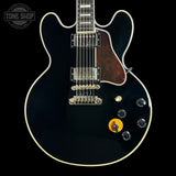 A black electric guitar with a glossy black finish, white binding, a dark brown pickguard, and gold hardware, shown against a solid black background with a Tone Shop watermark in the top left corner.