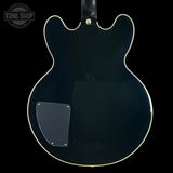 The back of a black electric guitar with a glossy finish, white binding, and a black pickguard, shown against a black background with a Tone Shop watermark in the top left corner.