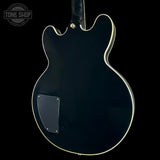 The back of a black electric guitar with a glossy finish, cream-colored binding, and a black pickguard, shown against a solid black background with a Tone Shop watermark in the top left corner.