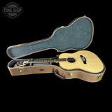 An acoustic guitar with a natural wood body and dark fretboard, shown inside an open hard case with a beige suede-like exterior and blue-gray interior padding, against a solid black background.