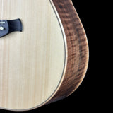 Close-up of an acoustic guitar body showing the light natural wood top, the dark brown figured wood side edge, and part of the black bridge against a black background.