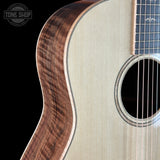 Close-up of an acoustic guitar showing the curved back panel with a rich brown wood grain, the light beige soundboard, and part of the dark fretboard, set against a black background with a Tone Shop watermark.