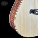 Close-up of an acoustic guitar body showing the light natural wood top, the darker wood edge, and part of the black soundhole surround against a black background. A Tone Shop watermark is visible in the upper left.