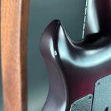Close-up of the side body of an electric bass guitar, showing a dark metallic purple finish with a small white scratch on the body, alongside a wooden stand and out-of-focus background.