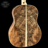Close-up of the back of an acoustic guitar showing a rich, figured wood grain pattern in warm brown and golden tones, with a light natural wood neck visible at the top against a solid black background.