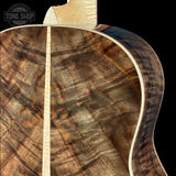 Close-up of the back of an acoustic guitar showing a figured wood top with a brown and tan grain pattern, a light center stripe, and a dark contoured side edge against a black background. A Tone Shop watermark is visible in the upper left.