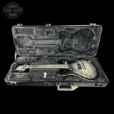An electric guitar with a black and gold flame finish sits inside an open black hard case with gray foam padding, set against a solid black background. A 'TONE SHOP' logo watermark is visible in the top left corner.