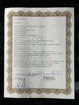A printed certificate or diploma with a decorative ornamental border, photographed from above on a dark surface. The document has cursive text, signatures, and a visible PDS logo near the upper right.