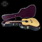 An acoustic guitar with a natural wood body and dark neck, displayed inside an open black hard case with a burgundy interior, set against a solid black background. A Tone Shop watermark is visible in the top-left corner.