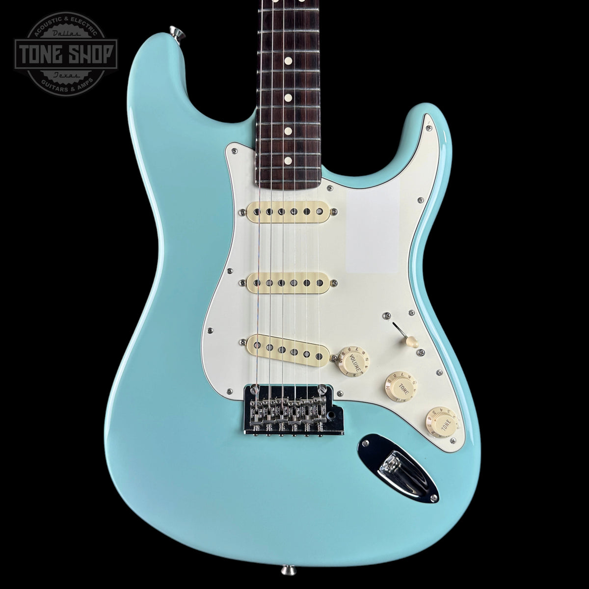 A light blue electric guitar with a white pickguard, cream-colored pickups and knobs, and a black bridge, shown against a solid black background. A Tone Shop watermark is visible in the top-left corner.