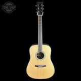 A natural wood acoustic guitar with a dark headstock, shown vertically against a solid black background. A circular 'TONE SHOP' watermark is visible in the top-left corner.