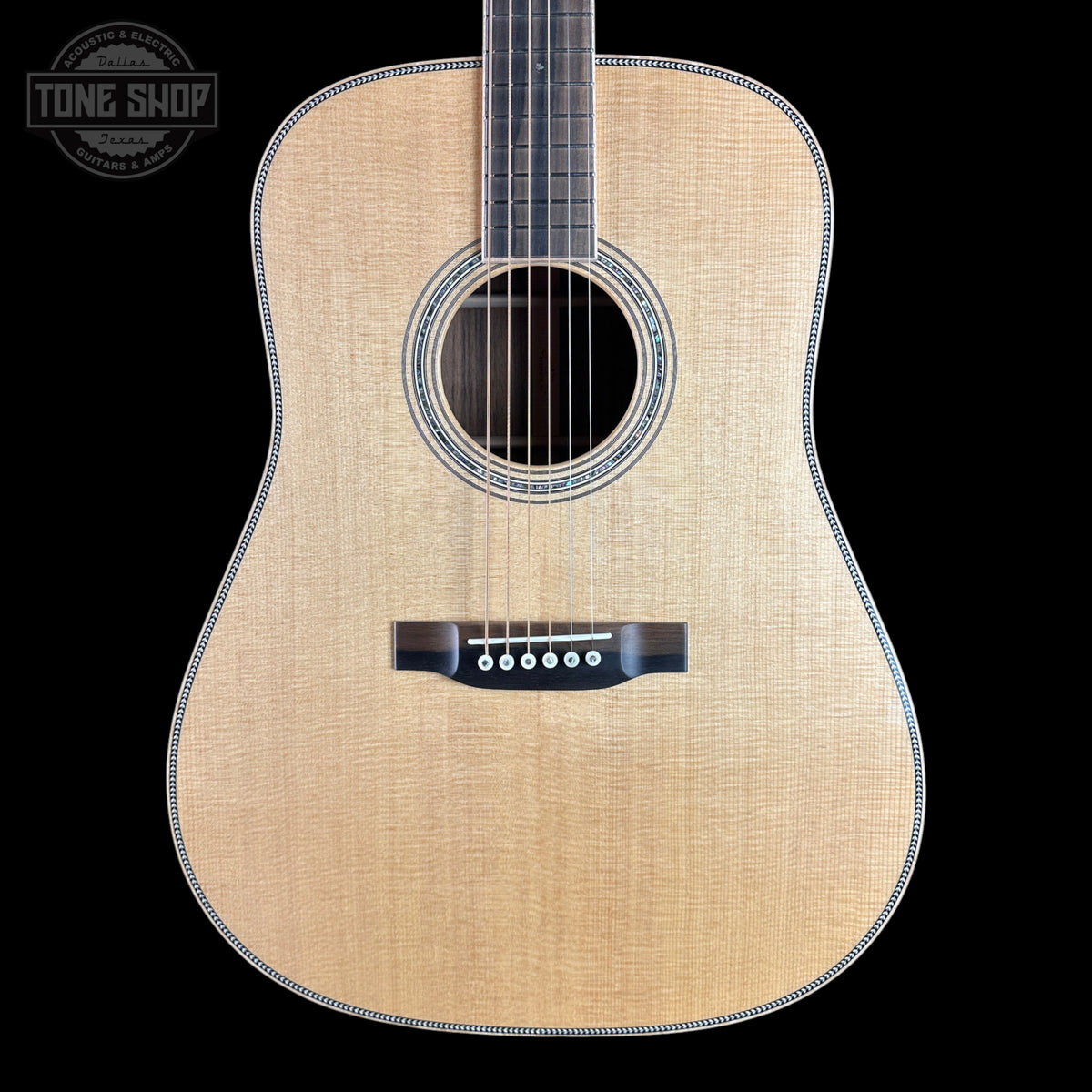 A front-facing acoustic guitar with a natural wood finish and black hardware, shown against a solid black background with a Tone Shop watermark in the top-left corner.