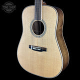 An acoustic guitar with a natural wood top and dark brown sides, shown against a solid black background with a Tone Shop watermark in the top-left corner.