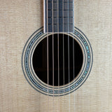 Close-up of an acoustic guitar body showing the round soundhole, rosette, strings, and part of the dark fretboard against a light wood grain background.