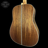 The back of a wooden acoustic guitar with a rich brown grain pattern and a light-colored binding, shown against a solid black background with a Tone Shop watermark in the top left.