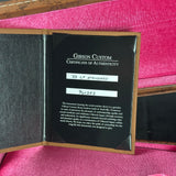 An open Gibson Custom Certificate of Authenticity booklet showing a black insert with white text and handwritten serial number labels, placed on a pink fabric surface.