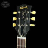 Close-up of a Gibson Les Paul Custom electric guitar headstock, featuring a black wood finish with gold 'Gibson' script, white 'Les Paul Custom' script, and six yellow tuning pegs against a black background.