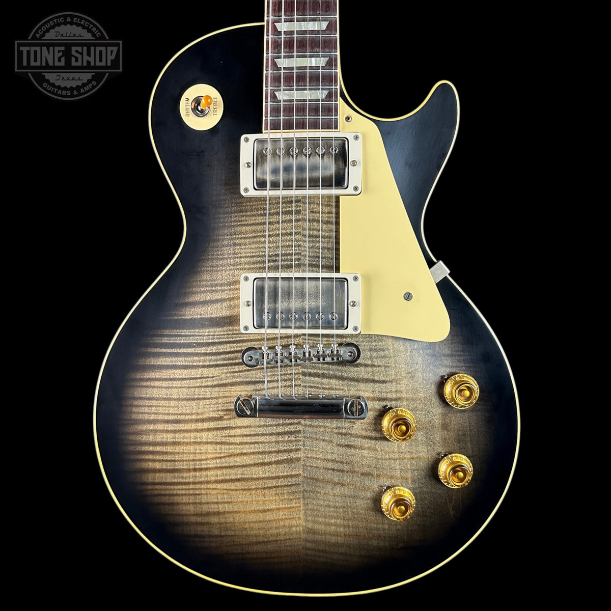A Gibson Les Paul Custom electric guitar with a natural flame maple top, featuring a black body finish, gold hardware, and a cream pickguard, shown against a solid black background.