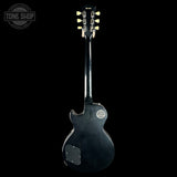 A black Gibson electric guitar shown from the back, featuring a glossy finish, six tuning pegs, and a circular Gibson badge on the body, set against a solid black background with a Tone Shop watermark in the top left.