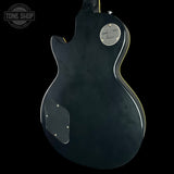 The back view of a black Gibson Les Paul Custom electric guitar against a solid black background, featuring a silver circular badge and a silver pickguard.